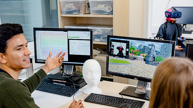 Zwei Forschende diskutieren vor ihren Rechnern. Im Hintergrund sitzt eine Probandin in einem kombinierten EEG-VR Experiment.