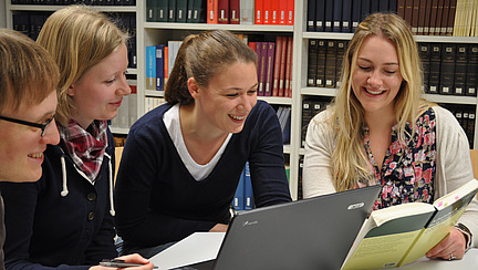 Four people in front of a wall of books looking at documents and a laptop together
