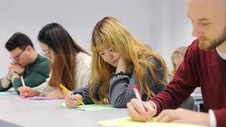 Vier Studierende an einem Tisch. Sie sehen konzentriert aus und schreiben auf buntem Papier.