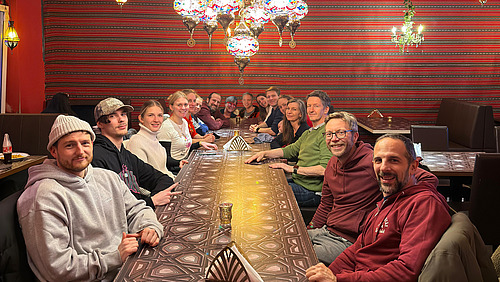 Kollegium sitzt an Tisch mit Kerzenleuchter &uuml;ber ihnen.