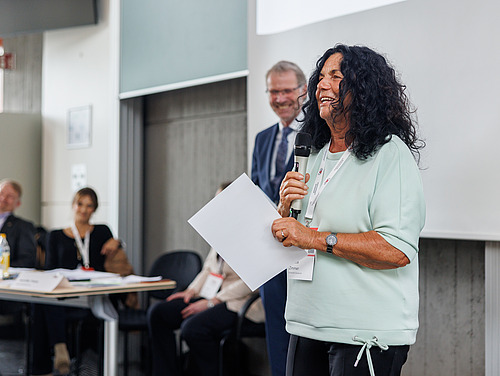 Eine Frau hält eine Urkunde und ein Mikrofon in den Händen und bedankt sich für eine Auszeichnung. 