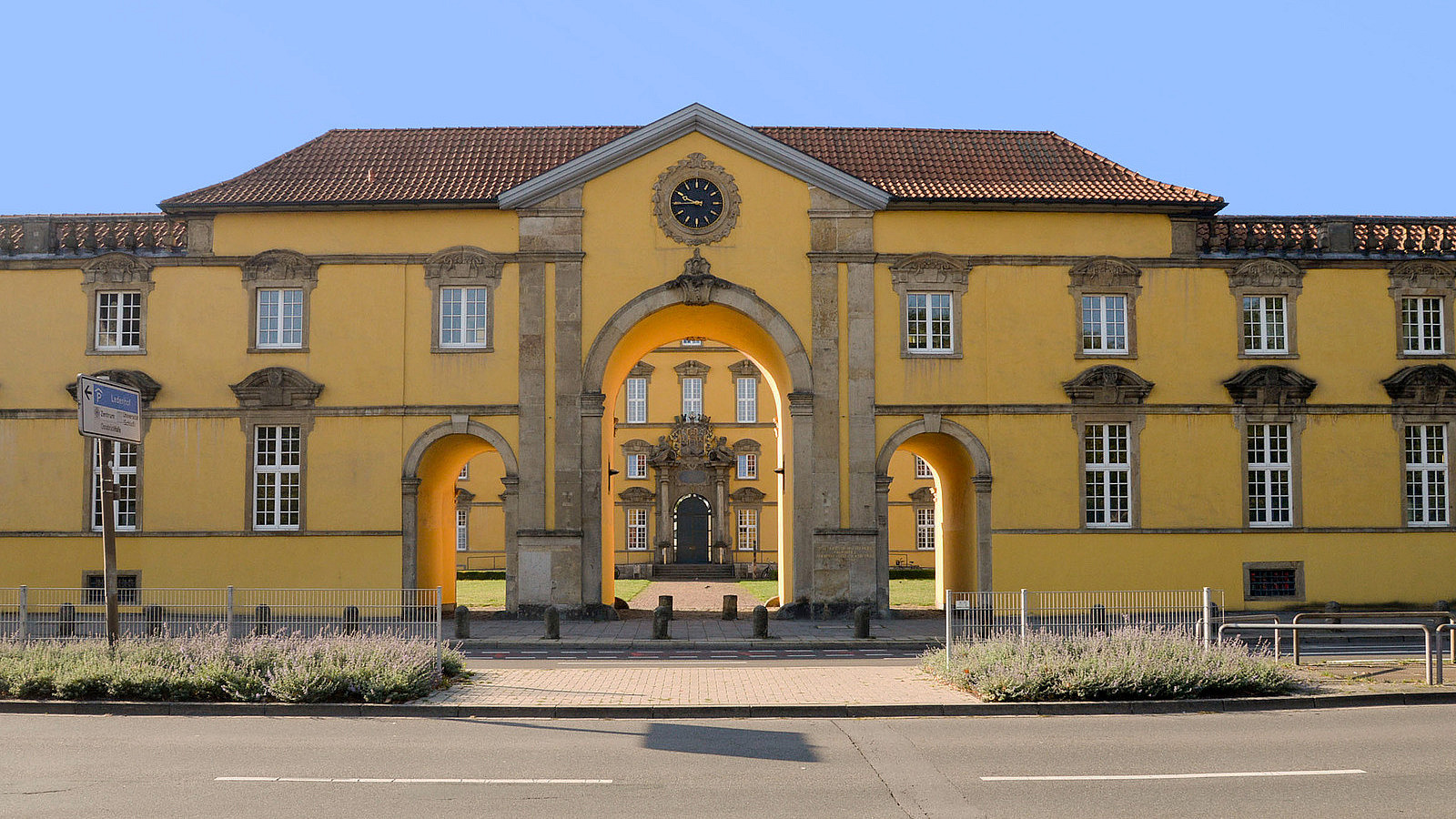 Das Haupttor des Schlosses über eine Straße fotografiert. Über dem Tor ist eine Uhr in die Mauer eingelassen.