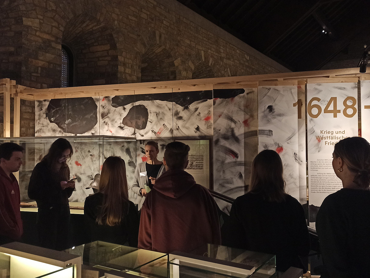 Das Bild zeigt eine Gruppe in einem Museum. Man sieht nur die Rücken. Im Hintergrund ist eine Tafel, auf der das Jahr 1648 steht.  