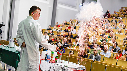 Die Weihnachtsvorlesung von Prof. Dr. Marco Beeken. Eine Person mit weißem Kittel hält einen Brenner an ein Reagenzglas. Aus dem Glas sprühen eine Menge Funken und Rauch.