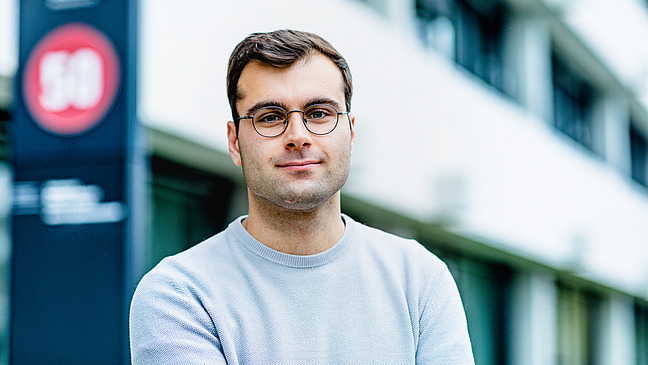 Das Bild zeigt Louis Loock vor dem Eingang des Instituts. Er lächelt mit verschränkten Armen in die Kamera.