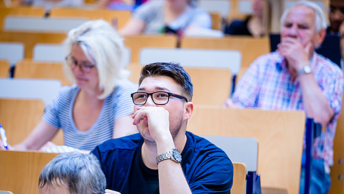 Blick über die Sitzreihen eines Hörsaals auf Studierende. Es sind drei ältere Menschen zu sehen.