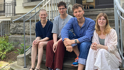 Vier junge Erwachsene sitzen auf einer Treppe. Im Hintergrund ist der Eingang zu einem öffentlichen Gebäude sichtbar