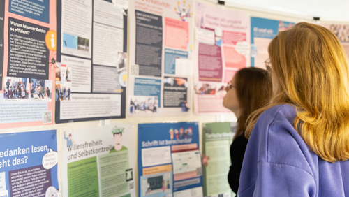 Zwei junge Frauen stehen vor einer Pinnwand, die mit bunten Plakaten bestückt ist
