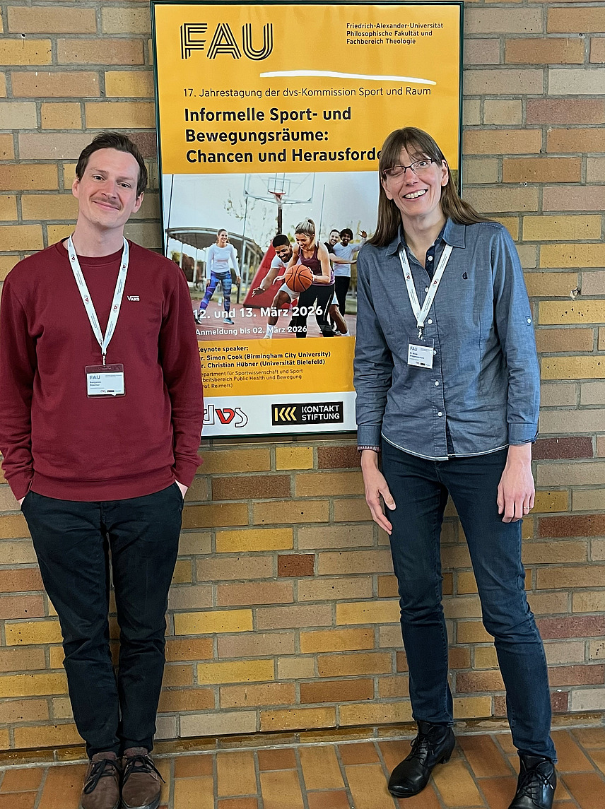 Herr Dr. des. Benjamin B&uuml;scher und Frau Dr. Br&uuml;ggemann-Kons stehen vor einem Plakat zum informellen Sport.