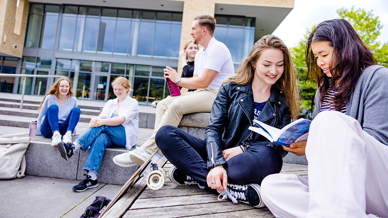 Eine Gruppe von Studierenden sitzt auf den Stufen vor einem modernen Universitätsgebäude und lernt oder entspannt sich.