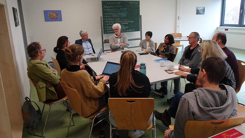 A group of 12 people is sitting around a table and discussing a topic. Two persons have their laptops open, one person is taking notes with a pen and paper. In the back we see a black board with notes written down.