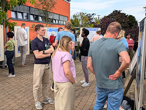 Personen stehen unter freiem Himmel vor Posterwänden mit wissenschaftlichen Postern und diskutieren.