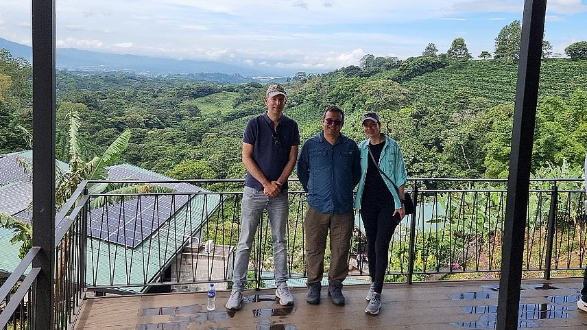 Das Bild zeigt eine malerische Aussicht von einem Aussichtspunkt aus, auf dem zwei Personen auf einer Holzplattform oder einem Balkon stehen. Man blickt auf eine &uuml;ppige Landschaft mit viel Gr&uuml;n und H&uuml;geln im Hintergrund. 