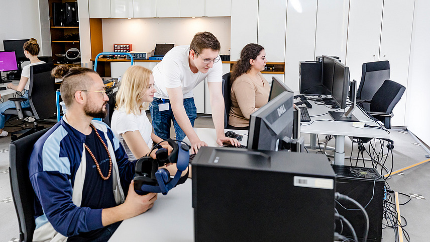 Vier Studierende sitzen an Computern während eine Person etwas am Computer erklärt. Sie befinden sich in den Räumlichkeiten des DFKI (Deutsches Forschungsinstitut für Künstliche Intelligenz).