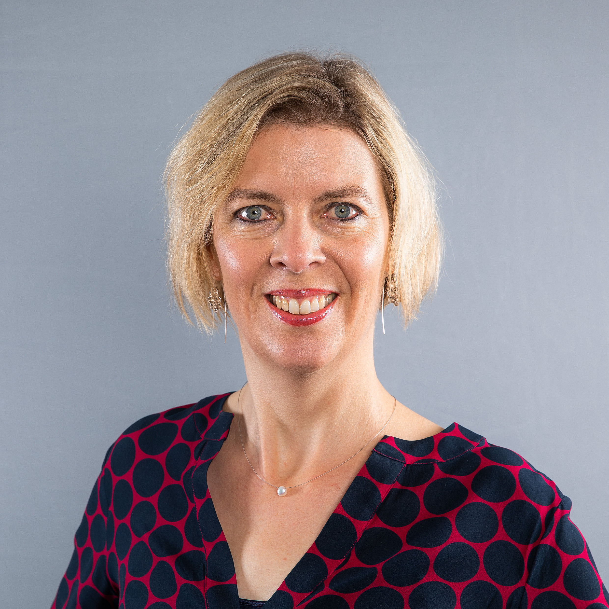 Eine lächelnde Frau mit kurzem, blondem Haar steht vor einem neutral grauen Hintergrund. Sie trägt eine dunkelblaue Bluse mit großem, roten Punktmuster sowie dezenten Schmuck – kleine Ohrringe und eine feine Halskette mit einem kleinen Anhänger. Die Frau schaut direkt in die Kamera.