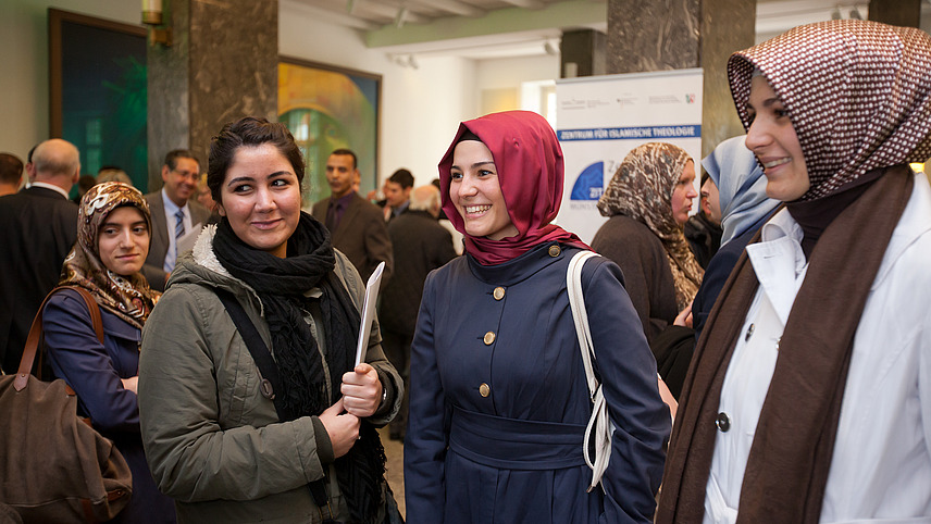 Four women in headscarves stand together in a semi-circle, smiling. In the background are other formally dressed people. 