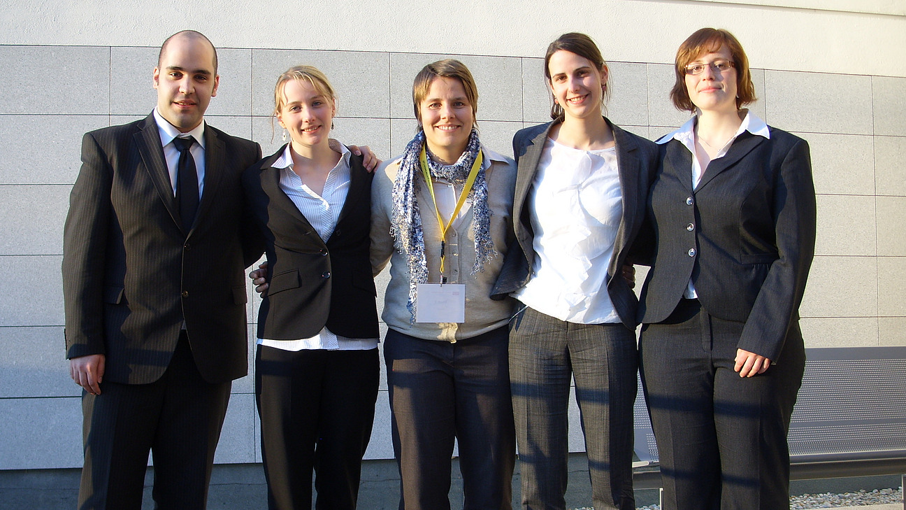 Ein Mann und vier Frauen in einer Reihe vor einer grauen Geb&auml;udewand stehend