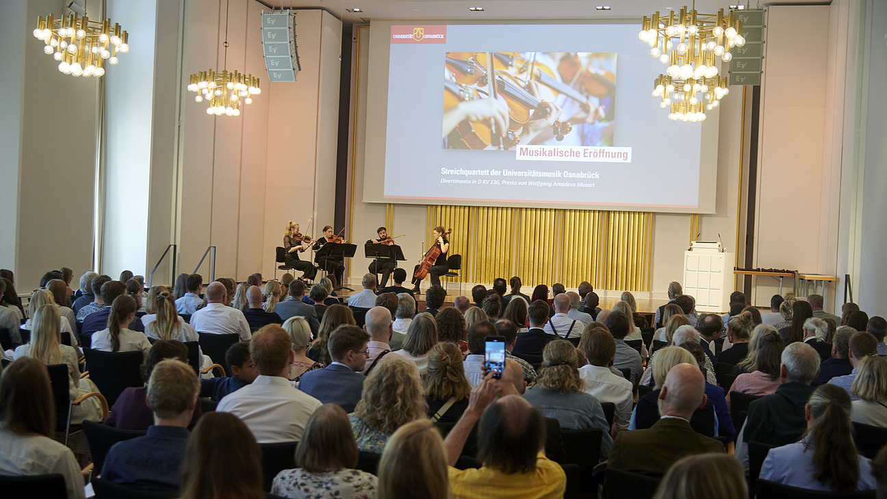 Vier Personen mit Streichinstrumenten auf einer Bühne. Im Publikum sitzen zahlreiche Personen