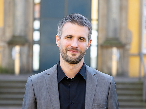 Mann mittleren Alters steht vor einem gro&szlig;en steinernen Geb&auml;ude und l&auml;chelt in die Kamera