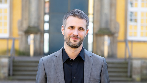 Mann mittleren Alters steht vor einem gro&szlig;en steinernen Geb&auml;ude und l&auml;chelt in die Kamera