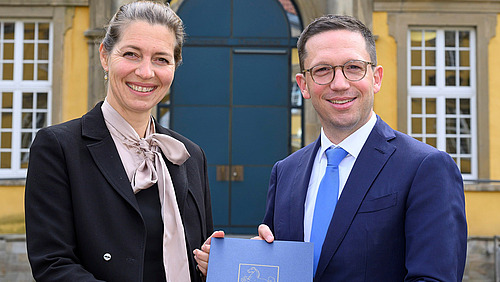 Susanne Menzel-Riedl und Falko Mohrs stehen vor dem Osnabrücker Schloss. Sie halten zusammen die Ernennungsurkunde in den Händen und lächeln in die Kamera.