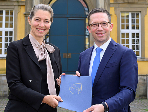 Susanne Menzel-Riedl und Falko Mohrs stehen vor dem Osnabrücker Schloss. Sie halten zusammen die Ernennungsurkunde in den Händen und lächeln in die Kamera.