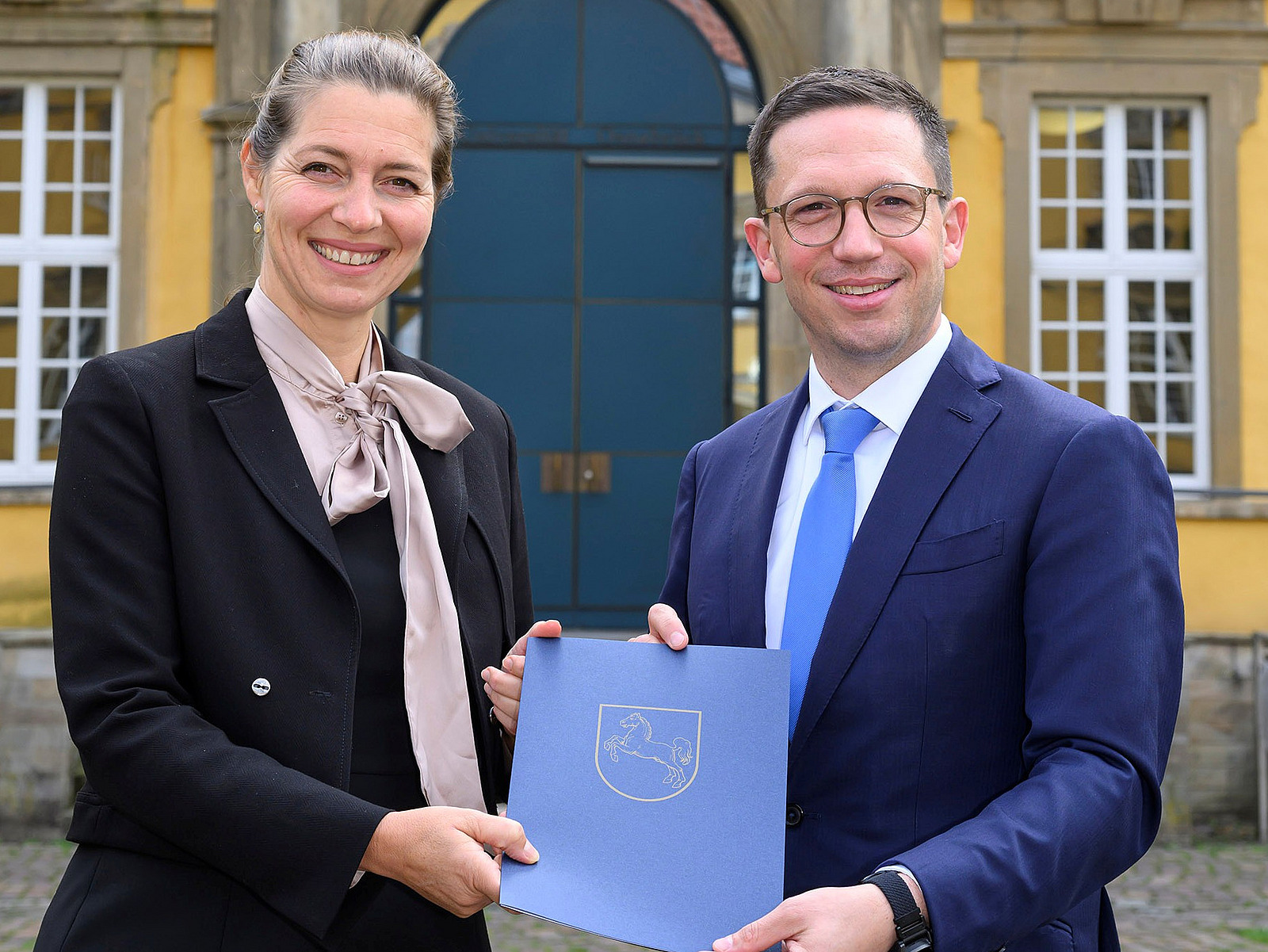 Susanne Menzel-Riedl und Falko Mohrs stehen vor dem Osnabrücker Schloss. Sie halten zusammen die Ernennungsurkunde in den Händen und lächeln in die Kamera.