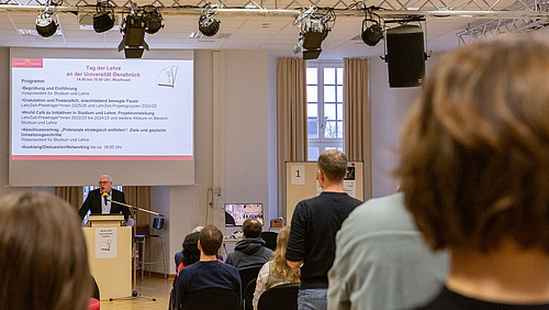 Ein Redner steht am Mikrofon und begrüßt Gäste in einem Saal.