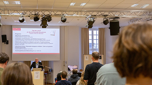 Ein Redner steht am Mikrofon und begrüßt Gäste in einem Saal.