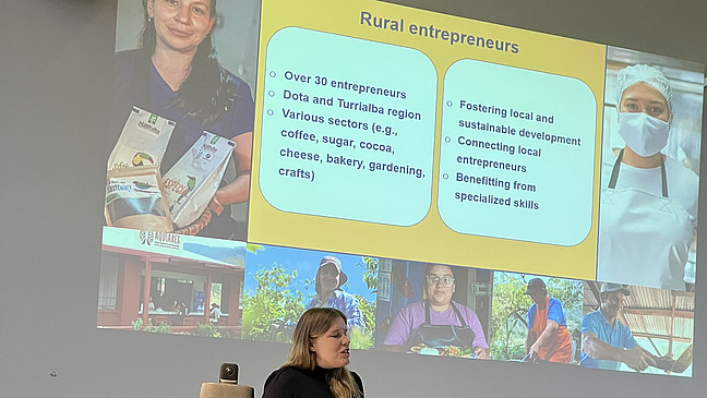 A person stands on a podium and gives a presentation to an audience about rural entrepreneurs in Costa Rica. The slide shows images of coffee and several people, assumingly working for the said entrepreneurs.