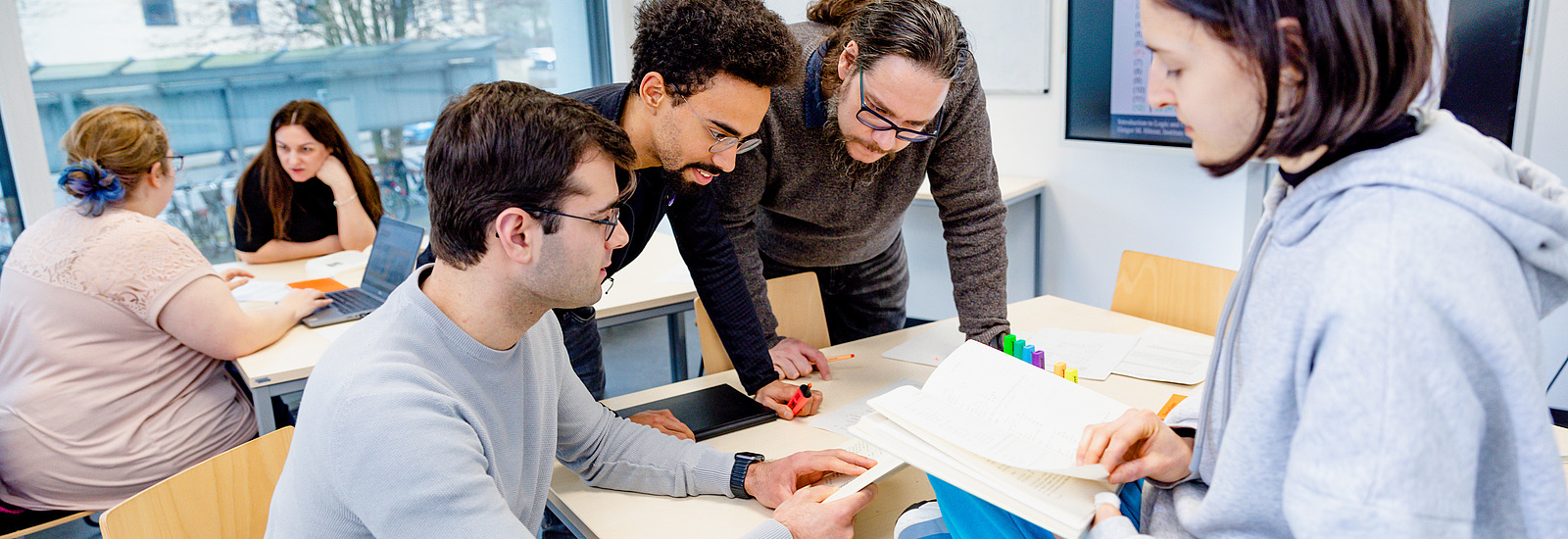 Eine Gruppe von drei Studierenden sitzt mit Studiendekan Gregor Hörzer an einem Tisch in einem modernen, gut beleuchteten Raum mit großen Fenstern und diskutieren etwas. Im Hintergrund sitzen zwei weitere Personen an einem anderen Tisch und diskutieren ebenfalls miteinander. 