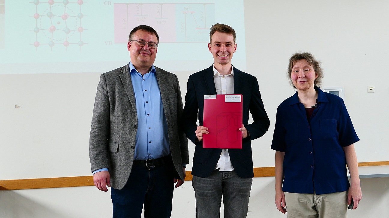 Ein Student mit Studiendekanin und Dekan stehen nebeneinander. Der Student hat eine Mappe in der Hand