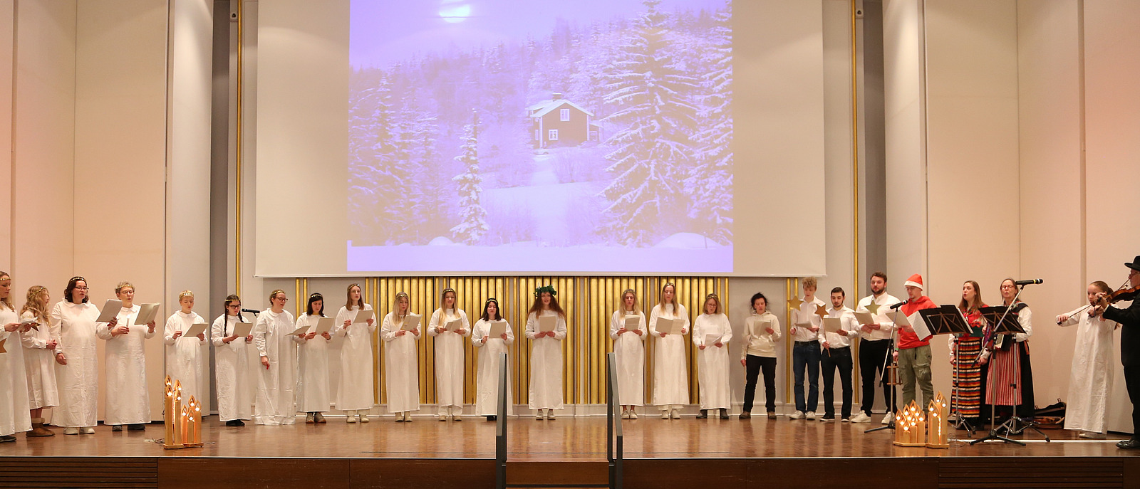 Der gesamte Chor singt auf der Bühne der Aula, begleitet vom Trio »Nymans Dans«. Auf die Leinwand ist eine Hütte im verschneiten Wald projiziert.