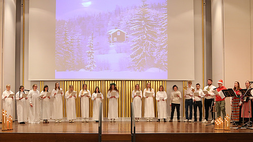 Der gesamte Chor singt auf der Bühne der Aula, begleitet vom Trio »Nymans Dans«. Auf die Leinwand ist eine Hütte im verschneiten Wald projiziert.