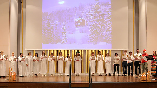 Der gesamte Chor singt auf der Bühne der Aula, begleitet vom Trio »Nymans Dans«. Auf die Leinwand ist eine Hütte im verschneiten Wald projiziert.
