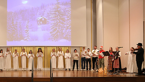 Der gesamte Chor singt auf der Bühne der Aula, begleitet vom Trio »Nymans Dans«. Auf die Leinwand ist eine Hütte im verschneiten Wald projiziert.