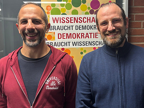 Benjamin Zander und Enrico Michelini stehen lächelnd vor einem Poster, auf welchems "Wissenschaft braucht Demokratie, Demokratie braucht Wissenschaft" geschrieben steht.
