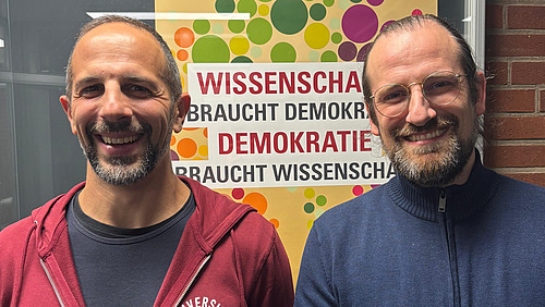 Benjamin Zander und Enrico Michelini stehen lächelnd vor einem Poster, auf welchems "Wissenschaft braucht Demokratie, Demokratie braucht Wissenschaft" geschrieben steht.