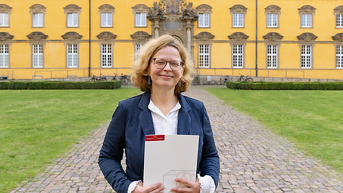 Portrait einer Frau mittleren Alters in Bluse und Blazer vor einem gro&szlig;en Geb&auml;ude mit vielen Fenstern