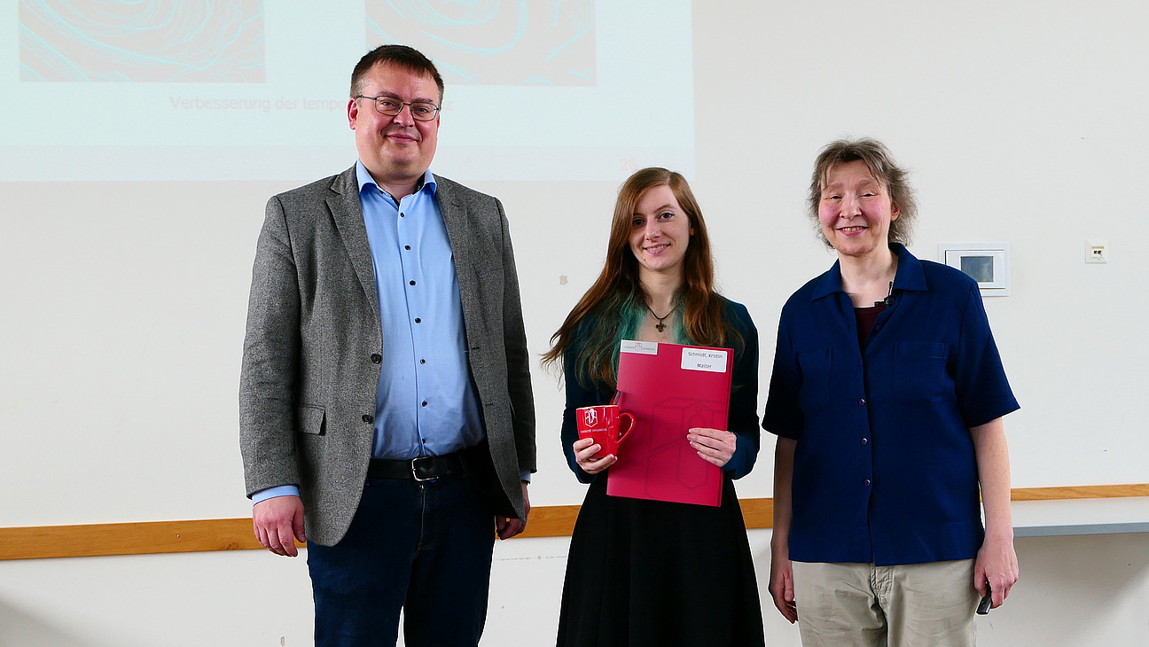 Eine Studentin mit Studiendekanin und Dekan stehen nebeneinander. Die Studentin hat eine Mappe in der Hand