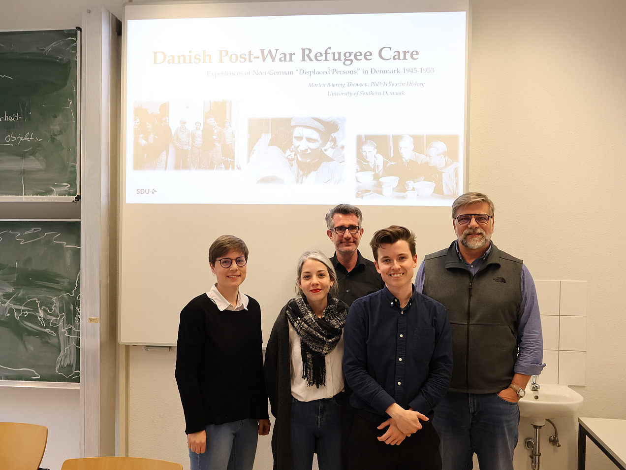 Das Foto zeigt fünf Personen, die in einem Raum vor einer Projektionsleinwand stehen. Auf der Leinwand ist der Titel "Danish Post-War Refugee Care" sichtbar. Im Hintergrund befindet sich eine Tafel mit handschriftlichen Notizen.