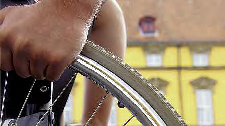 Zwei Reifen eines Rollstuhls werden von Händen gegriffen, im Hintergrund ist ein großes gelbes Gebäude.