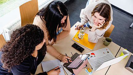 Drei Frauen sitzen zusammen an einem Tisch und arbeiten mit einem Laptop, einem Tablet, Papier und Stift. Die Szene ist von oben zu sehen und ist hell erleuchtet.