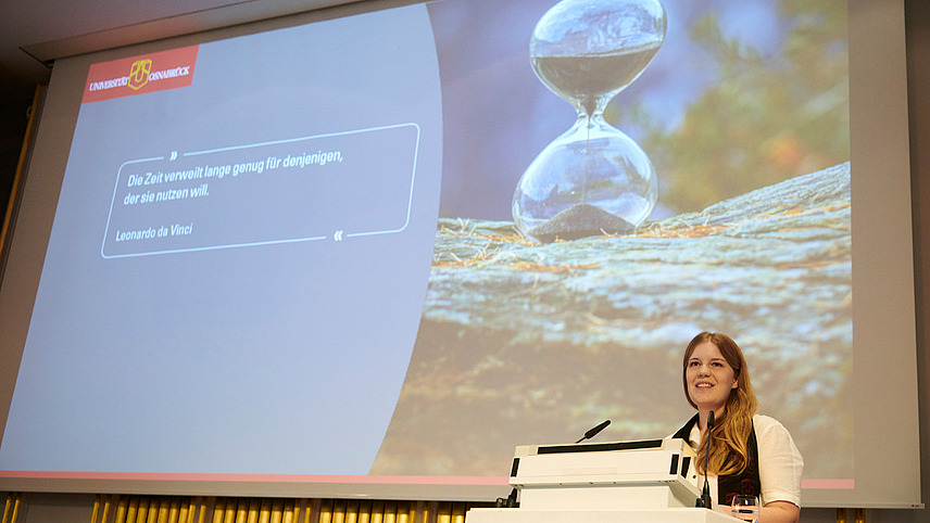 A person stands at a lectern, in the background you can see a presentation slide with a large hourglass and a quote from Leonardo da Vinci: "Time lingers long enough for those who want to use it.