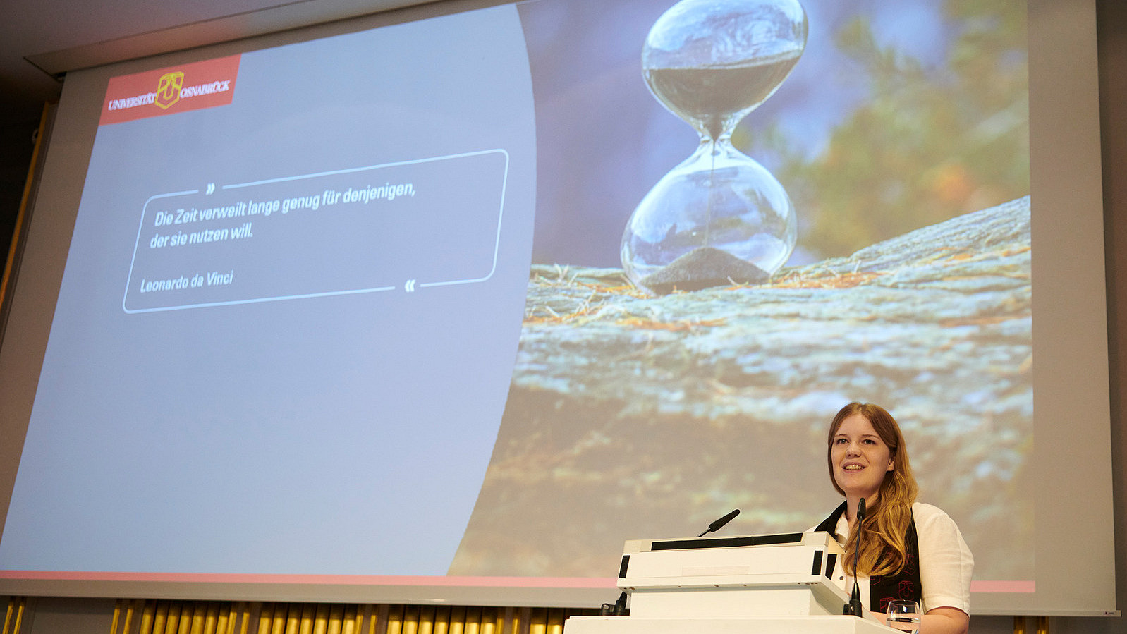 A person stands at a lectern, in the background you can see a presentation slide with a large hourglass and a quote from Leonardo da Vinci: "Time lingers long enough for those who want to use it.