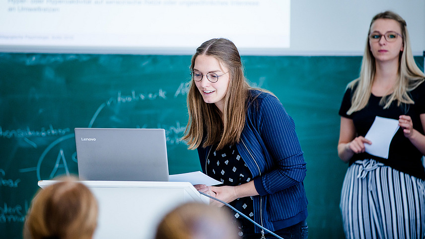 A student in a seminar room is standing in front of a laptop on a desk. She is looking at the laptop. A blackboard can be seen behind her.