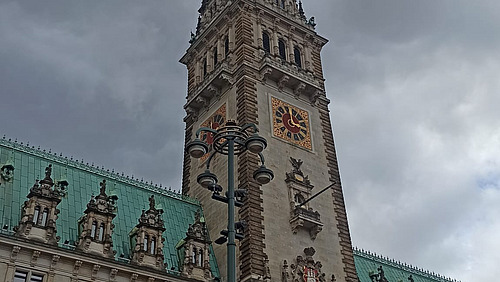 Das Bild zeigt das Hamburger Rathaus. Es ist ein beigefarbenes Geb&auml;ude mit einem hohen hohen Turm in der Mitte. Das Dach ist t&uuml;rkis.