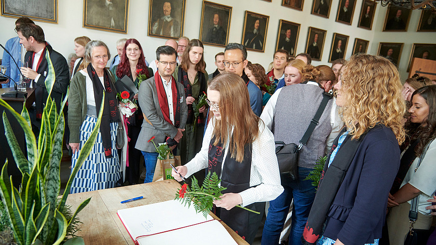In a historic-looking building, several people are standing at a table on which an open book with blank pages is lying. One person is writing something in the book.