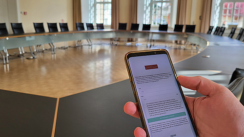 Konferenzsaal ohne Personen, im Vordergrund hält eine Hand ein Smartphone ins Bild
