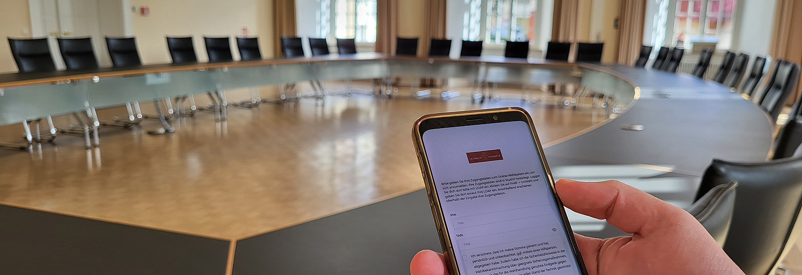 Konferenzsaal ohne Personen, im Vordergrund hält eine Hand ein Smartphone ins Bild